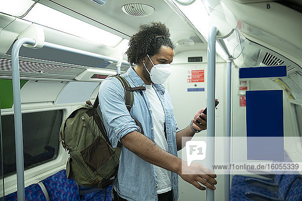 Mann mit Schutzmaske steht im U-Bahn-Zug und schaut auf sein Handy  London  UK