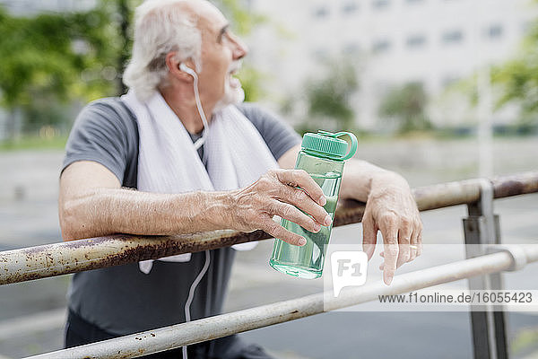 Älterer Mann hält eine Flasche und hört Musik  während er an einem Geländer in der Stadt steht