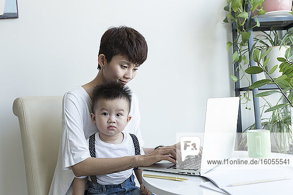Mutter mit süßem Jungen auf dem Schoß arbeitet über Laptop zu Hause