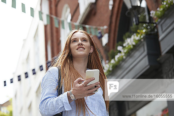 Lächelnde rothaarige Frau schaut weg  während sie mit ihrem Smartphone gegen ein Gebäude in der Stadt läuft