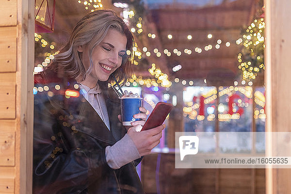 Lächelnde Frau mit heißer Schokolade und Smartphone in einem Geschäft im Vergnügungspark  gesehen durch ein Fenster