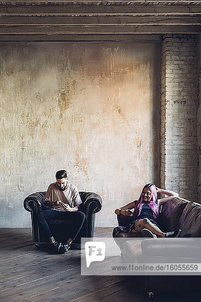 Junger Mann und Frau sitzen auf Sofa und Sessel in einem Loft und benutzen Smartphones