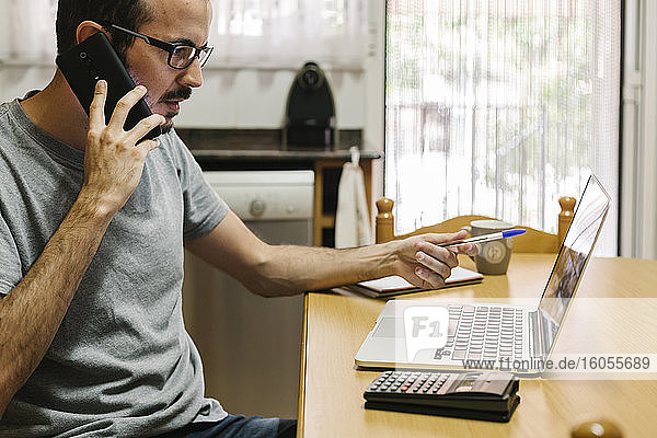 Mittelgroßer erwachsener Mann  der ein Smartphone benutzt  während er zu Hause am Laptop arbeitet