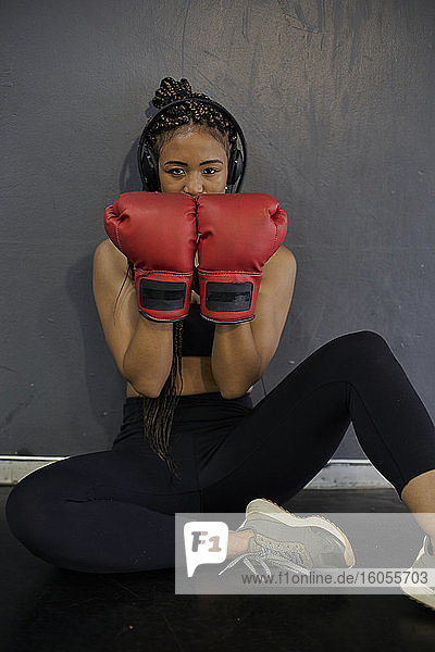 Junge Frau mit roten Handschuhen hört Musik über Kopfhörer  während sie sich in einem Fitnessstudio an die Wand lehnt