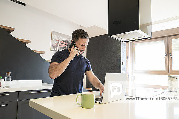 Mann  der über sein Smartphone spricht  während er einen Laptop auf der Kücheninsel zu Hause benutzt