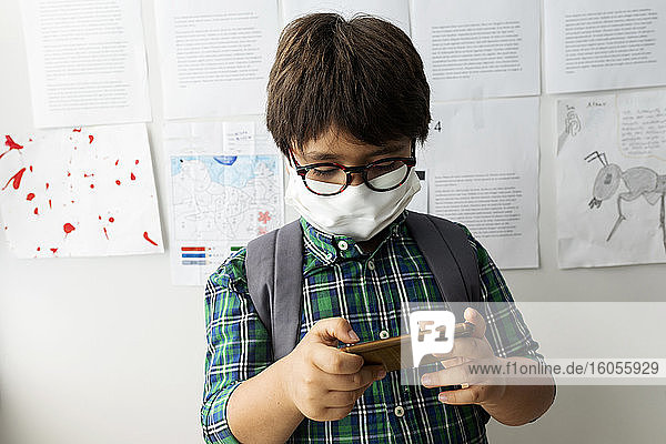 Junge mit Maske  der ein Mobiltelefon benutzt  während er in der Schule an der Wand steht