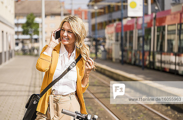Glückliche Geschäftsfrau  die ihr Smartphone benutzt  während sie auf dem Gehweg neben der Straßenbahn steht