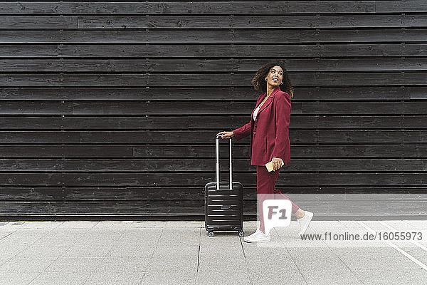 Young businesswoman looking back while walking with luggage on footpath by black wooden wall in city