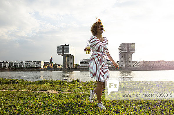Cheerful woman with beer bottle walking on grassy land against river in city at sunset