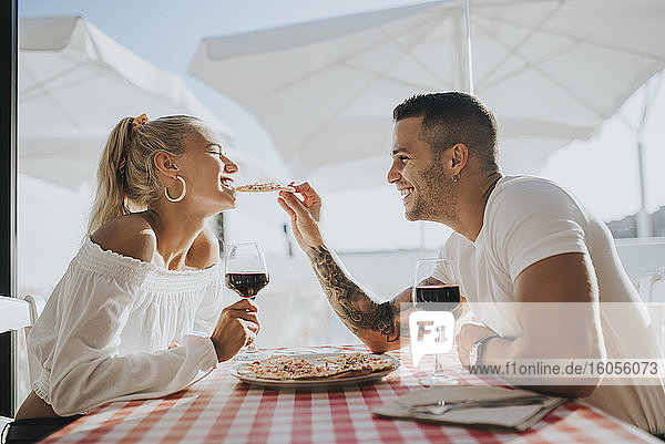 Happy young man feeding pizza to girlfriend while sitting in restaurant