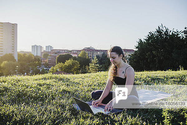 Sportliche junge Frau benutzt einen Laptop  während sie auf einer Übungsmatte im Stadtpark sitzt