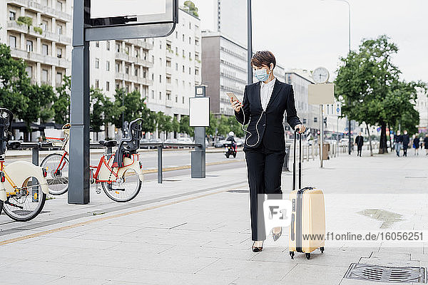 Geschäftsfrau mit Maske  die ein Smartphone benutzt  während sie mit einem Koffer auf dem Gehweg in der Stadt spazieren geht