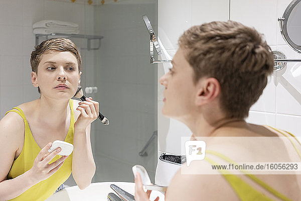 Schöne Frau mit kurzen Haaren trägt Make-up auf und betrachtet ihr Spiegelbild im Badezimmer