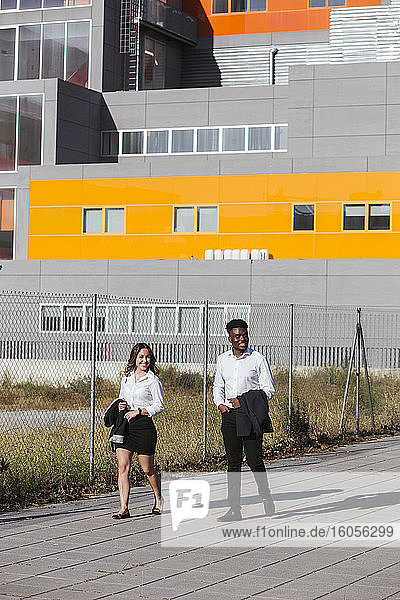 Lächelnde Kollegen  die an einem sonnigen Tag auf einem Fußweg vor einem modernen Gebäude spazieren gehen