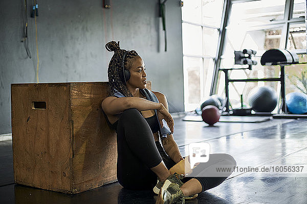 Nachdenkliche Sportlerin hört Musik  während sie im Fitnessstudio auf dem Boden sitzt