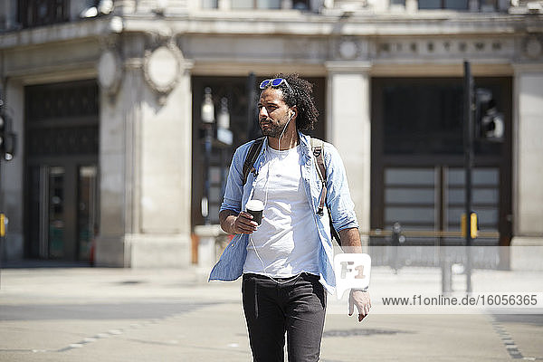 Portrait of young man with coffee to go crossing the street while listening music with earphones  London  UK
