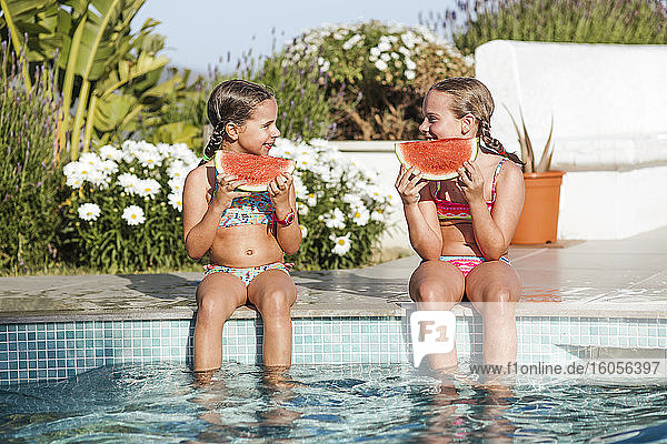 Lächelnde Zwillingsmädchen sitzen mit Wassermelonenscheibe am Pool an einem sonnigen Tag