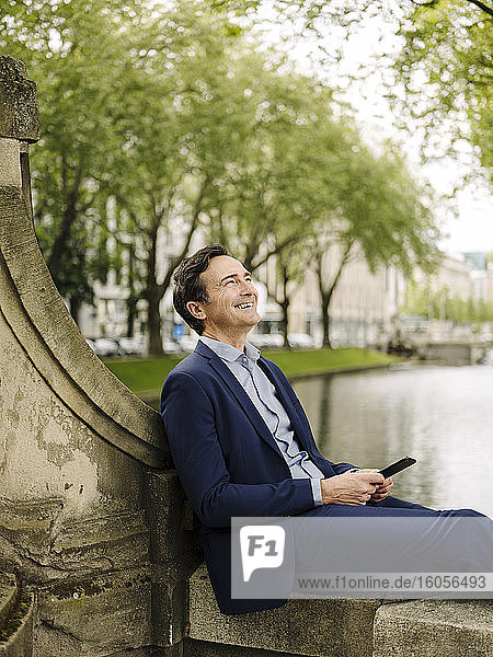 Glücklicher reifer Geschäftsmann mit Smartphone  der auf einer Brückenbrüstung sitzt