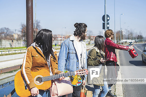Young man hitchhiking while standing with friends on street in city during sunny day