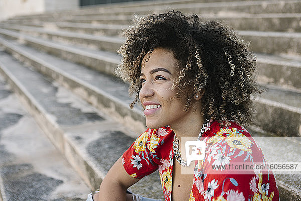 Nachdenkliche junge Frau lächelt  während sie auf den Stufen eines sonnigen Tages sitzt