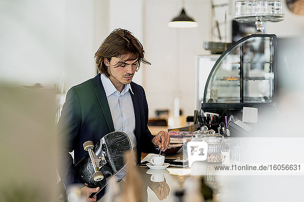 Businessman holding skateboard while mixing coffee on counter in cafe