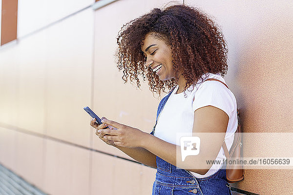 Glückliche junge Frau mit Afro-Haar  die ein Smartphone benutzt  während sie an einer Mauer in der Stadt steht