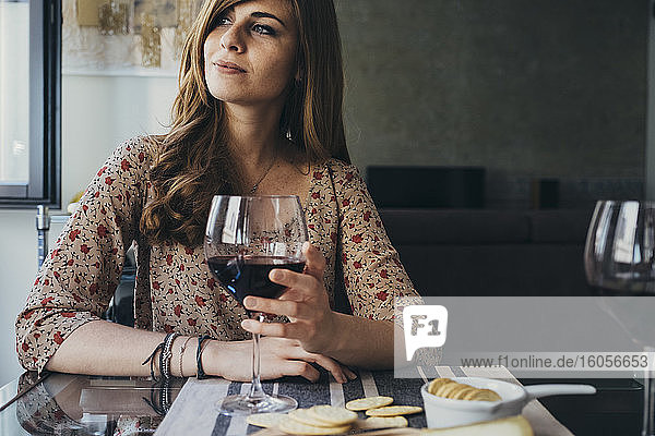 Beautiful young woman holding wineglass looking away while sitting at dining table