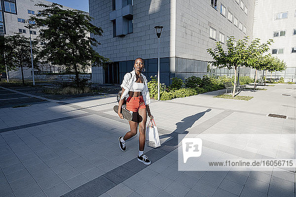 Junge Frau mit Einkaufstasche und Skateboard auf dem Fußweg in der Stadt
