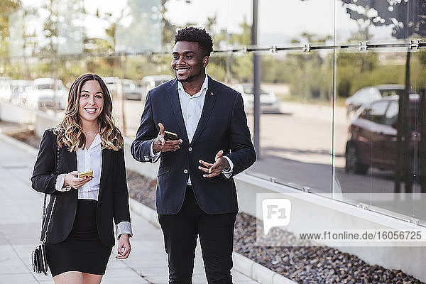 Lächelnde Geschäftsleute  die ihr Smartphone in der Hand halten  während sie auf dem Gehweg in der Stadt spazieren gehen