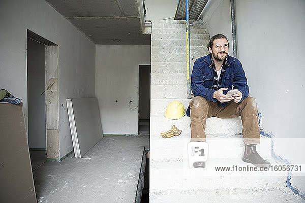 Nachdenklicher Bauarbeiter hält sein Smartphone in der Hand  während er auf den Stufen eines im Bau befindlichen Hauses sitzt
