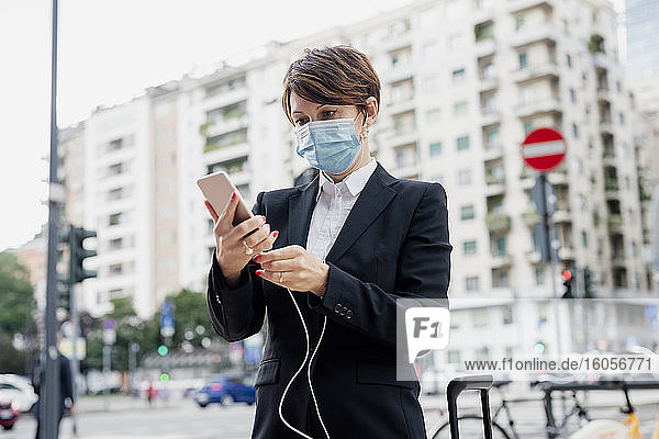 Businesswoman wearing mask holding smart phone and headphones while standing in city