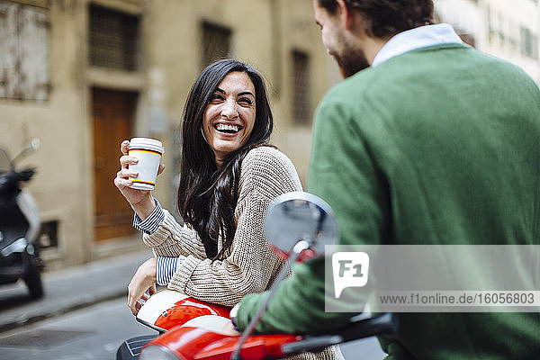 Glückliche Frau mit Kaffee in der Hand  die ihren Freund anschaut  während sie sich auf eine Vespa lehnt