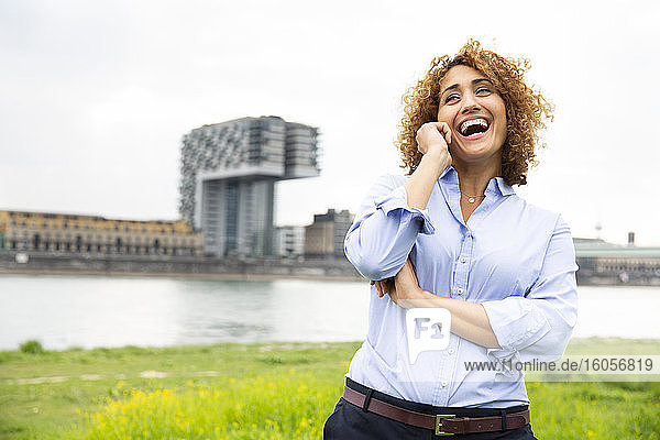 Fröhliche Geschäftsfrau mit lockigem Haar  die über ein Mobiltelefon spricht  während sie gegen den Himmel in der Stadt steht