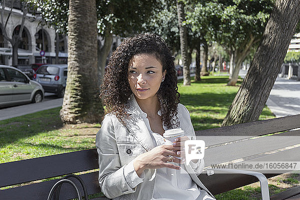 Nachdenkliche  stilvolle Geschäftsfrau hält eine Kaffeetasse  während sie auf einer Bank im Park sitzt