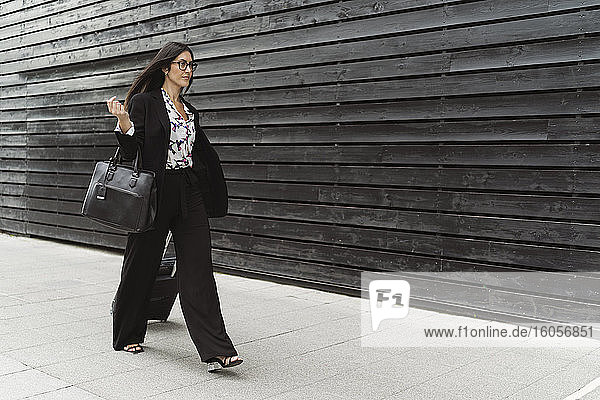 Selbstbewusste junge Geschäftsfrau mit Gepäck auf dem Fußweg an der schwarzen Wand