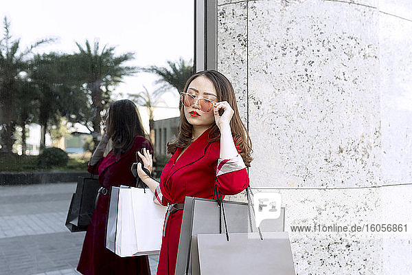 Junge Frau mit Sonnenbrille trägt Einkaufstaschen  während sie an einem modernen Gebäude steht