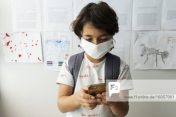 Junge mit Maske benutzt sein Smartphone  während er in der Schule an der Wand steht