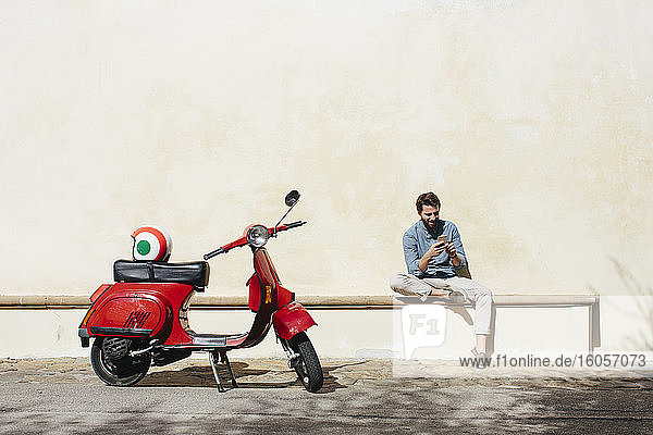 Young men using smart phone while sitting on seat against wall