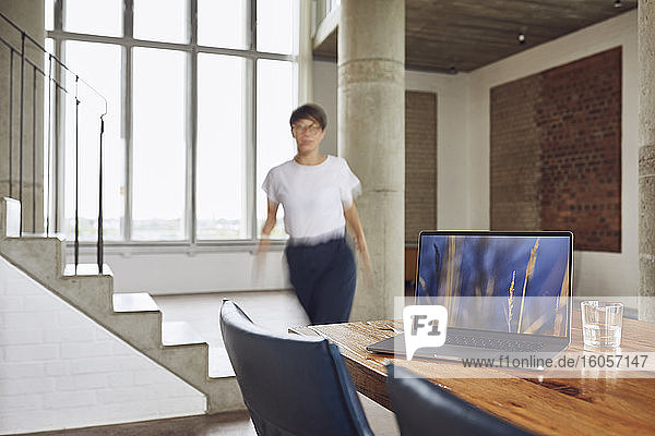 Laptop auf Holztisch in einer Loftwohnung mit Frau im Hintergrund