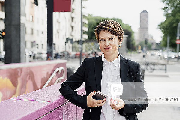 Lächelnde Geschäftsfrau  die ihr Smartphone und ihre Kopfhörer hält  während sie an einer Stützmauer in der Stadt steht