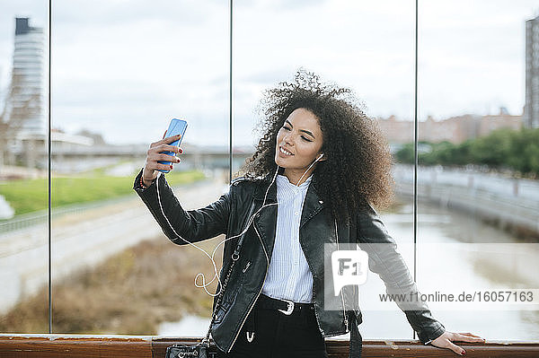 Lächelnde junge Afro-Frau  die ein Selfie macht  während sie sich auf ein Geländer stützt