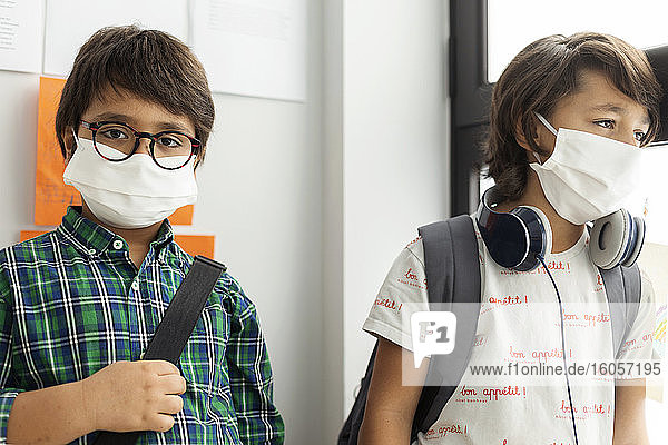 Junge mit Maske  der mit einem Freund an der Wand einer Schule steht