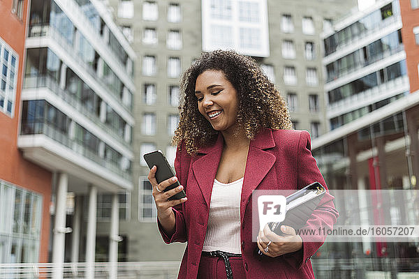Smiling young businesswoman using smart phone while standing against building in city