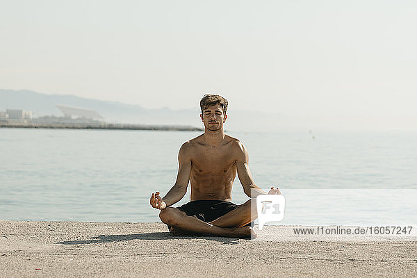 Hemdloser junger Mann meditiert im Sitzen gegen das Meer am Hafen