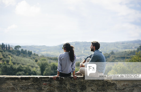 Paar betrachtet die Landschaft  während es auf einer Stützmauer gegen den Himmel sitzt