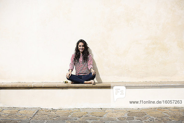 Smiling young woman sitting on seat against wall during sunny day