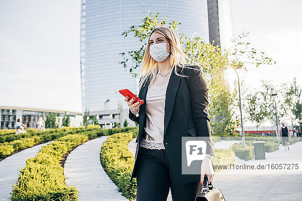 Businesswoman wearing face mask holding smart phone while walking on footpath in city