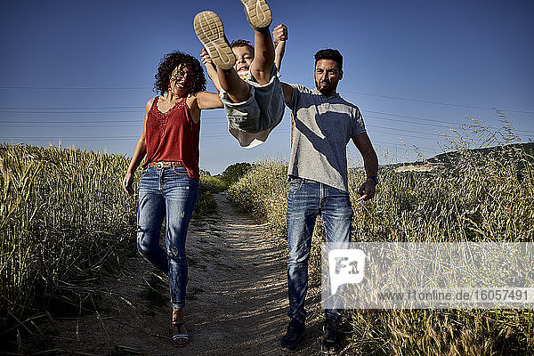 Boy jumping while holding parents hands in farm against clear blue sky
