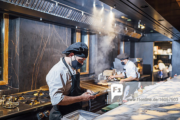 Chef wearing protective face mask using smartphone in restaurant kitchen