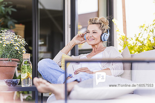 Porträt einer lächelnden reifen Frau  die auf einer Terrasse sitzt und mit Kopfhörern Musik hört